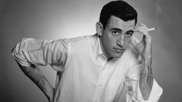 Fotografía en blanco y negro de J. D. Salinger de joven, con un cigarrillo en la mano con la que se toca la frente. La otra mano está apoyada en su cadera. Viste una camisa blanca.