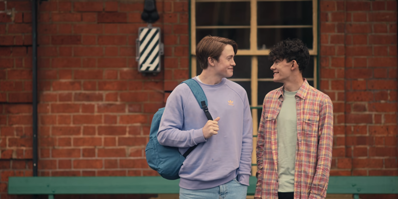 Fotograma de Nick y Charlie en una estación de tren. Nick viste una sudadera lila y Nick una camiseta gris y una camisa de cuadros roja. Se están mirando y sonriendo.